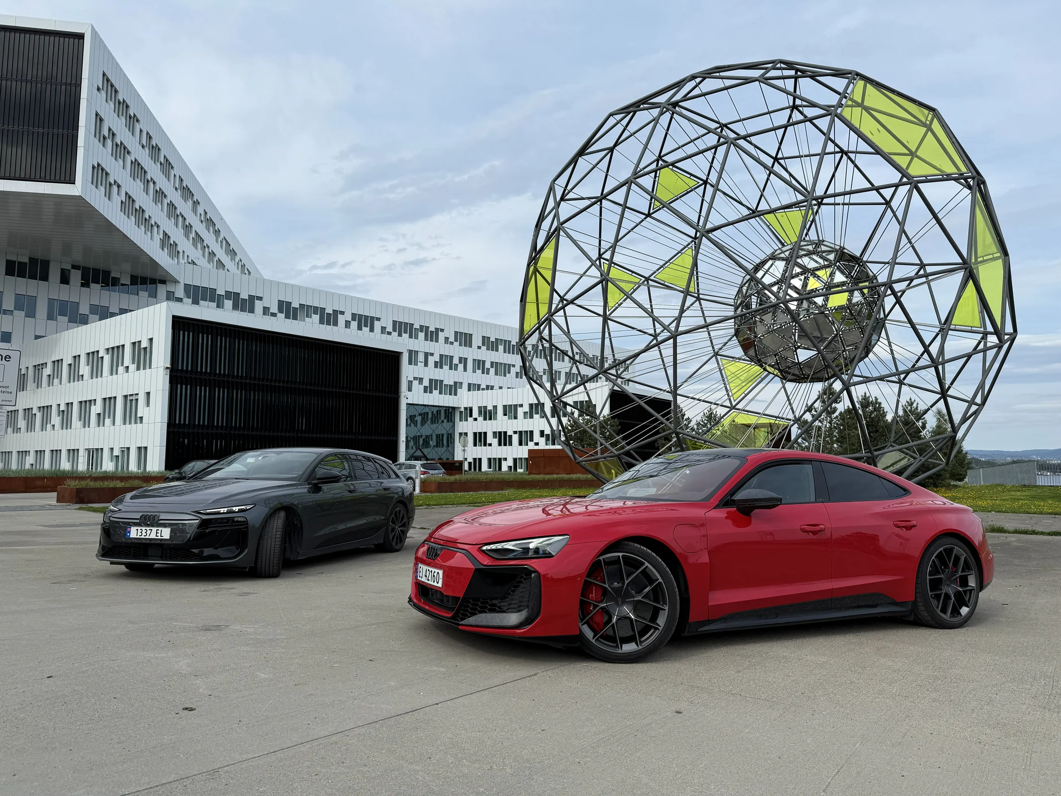 A dark Audi S6 e-tron Avant and a red Audi RS e-tron GT performance in front of a large geometric metal sculpture.